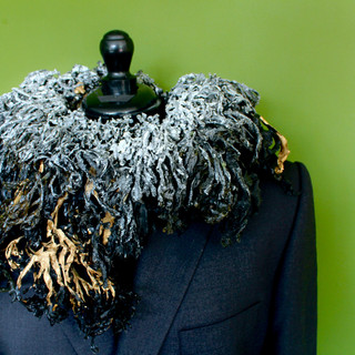 A pale-skinned man stands facing left against a green background. You can see from his nose to his upper chest. He is bare chested and wears warrior's ruff made of dried, black seaweed around his neck. The seaweed is thick and deep and looks like thousands of black feathers. The seaweed is covered in a heavy salt crust just around the neck, some of which forms large crystals. As the seaweed moves away from the neck, it gets blacker and shinier. At the very front at the bottom, is is gilt with gold paint.