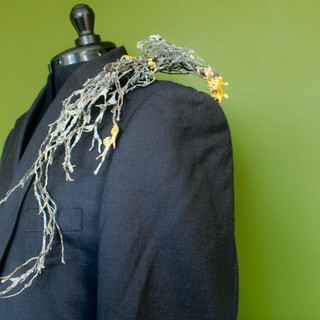 A grey suit jacket on a dressmaker's dummy  stands facing left against a green background. Resting on the left shoulder is a wave boa made of dried, black seaweed. The seaweed appears to rush from the neck down the front of the jacket. The seaweed is covered in a heavy salt crust, some of which form large crystals. At a few points on the boa, the seaweed is is gilt with fluttering gold leaf. 