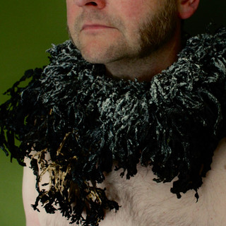 A pale-skinned man stands facing left against a green background. You can see from his nose to his upper chest. He is bare chested and wears a warrior's ruff made of dried, black seaweed around his neck. The seaweed is thick and deep and looks like thousands of black feathers. The seaweed is covered in a heavy salt crust just around the neck, some of which form large crystals. As the seaweed moves away from the neck, it gets blacker and shinier. At the very front at the bottom, is is gilt with gold paint.