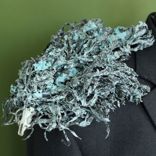 A grey suit jacket on a dressmaker's dummy  stands facing out against a green background. resting around the jacket's neck is a warrior's ruff made of dried, black seaweed. The seaweed is thick and deep and looks like thousands of black feathers cascading out. The seaweed is covered in a heavy salt crust just around the neck, some of which form large crystals. As the seaweed moves away from the neck, it gets blacker and shinier. At the very front at the bottom of the ruff, and just on the left shoulder, the seaweed is is gilt with gold paint.