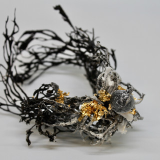 A bracelet made of dried, black seaweed, salt crust and gold leaf sits on a white background. The seaweed curls around to make a bracelet shape with tendrils reaching from it. The black seaweed has a light dusting of salt all over it. On the top of the bracelet, sits a corsage. The corsage is made of salt crystals, salt-encrusted seaweed bladders and fluttering gold leaf. 