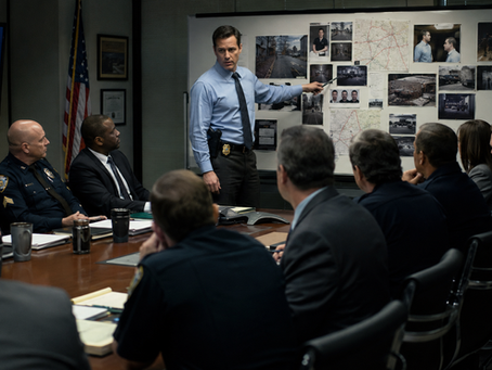 Detective briefs police and officials in a conference room, pointing to photos and maps on a homicide investigation board
