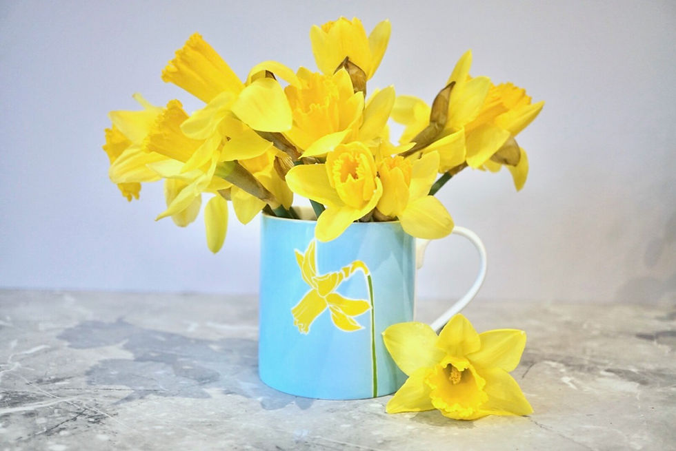 A mug featuring a yellow daffodil flower on a blue background, with fresh yellow daffodils inside on a grey countertop