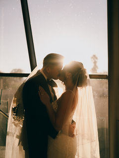 A couple in wedding attire kisses in front of a sunlit window. The bride's veil glows in the soft sunlight, creating a romantic atmosphere.