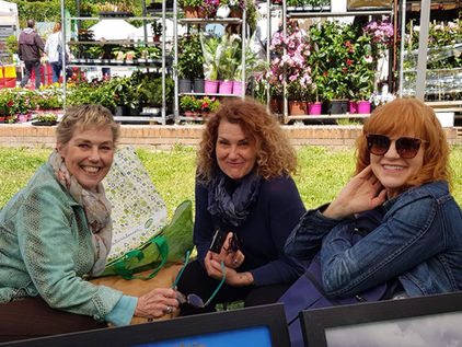 Three smiling women sitting on grass at an outdoor flower market.