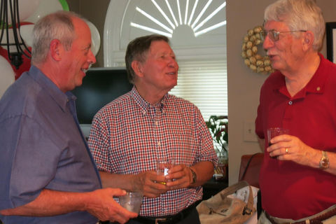 Three men smiling, holding drinks, and conversing at a social gathering.