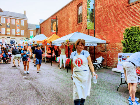 Smiling woman in apron at bustling outdoor market, 'DRINK TICKETS' tent.