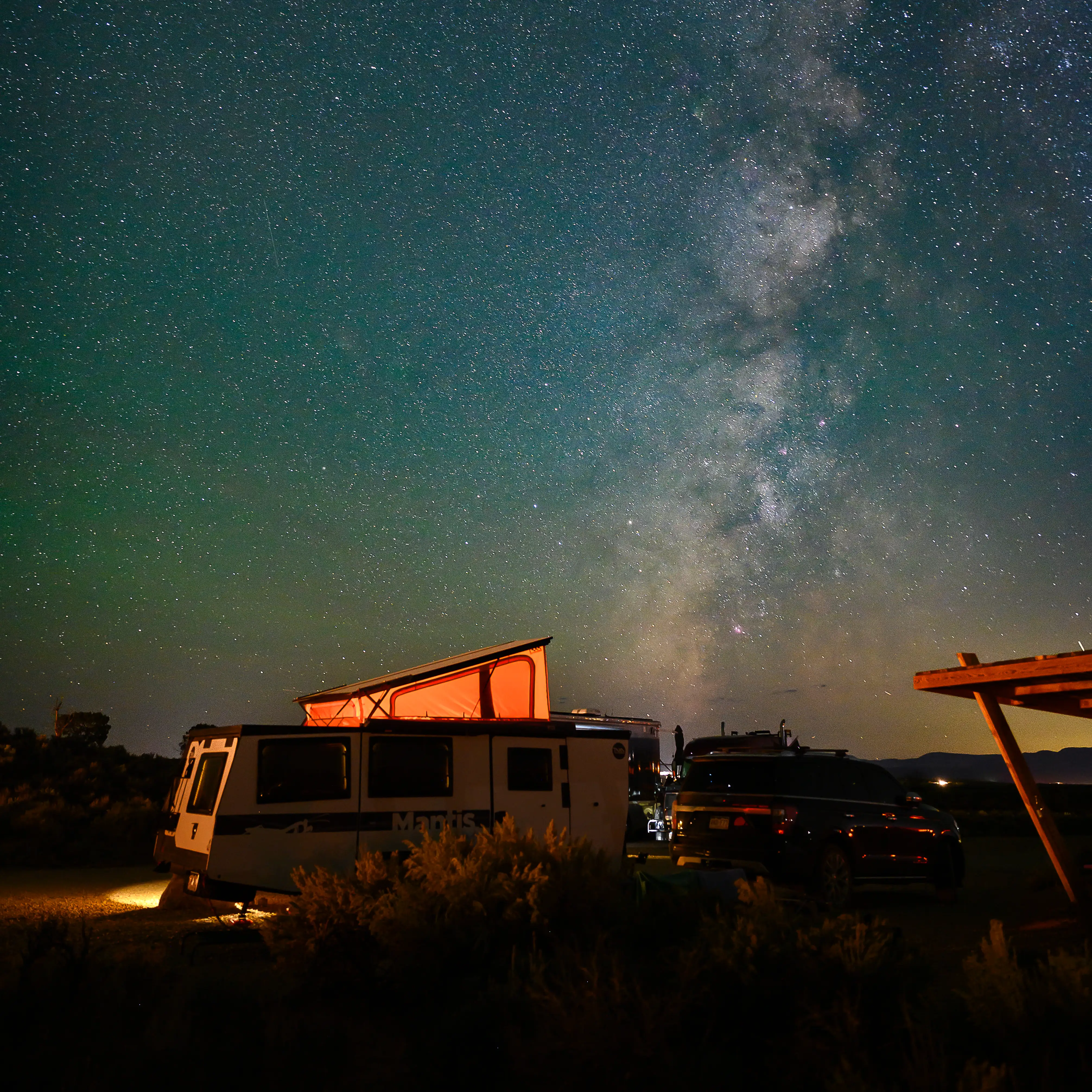 Nighttime camping scene in Kanab, Utah featuring a glowing camper trailer under a star-filled sky with the Milky Way galaxy stretching overhead.