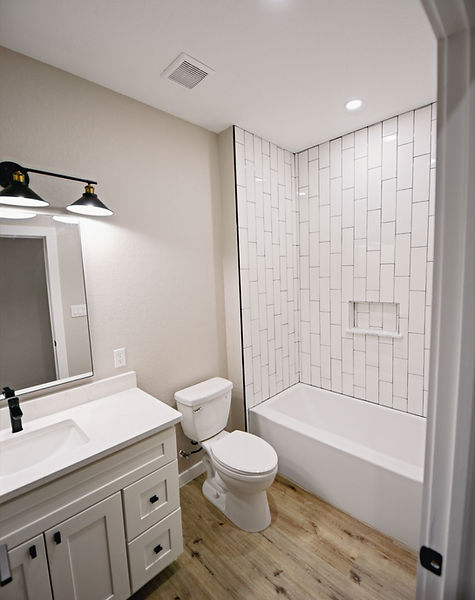 First of two bathrooms with crisp white tile, cabinetry and fixtures with black hardware and grout accenting oak colored luxury vinyl plank flooring