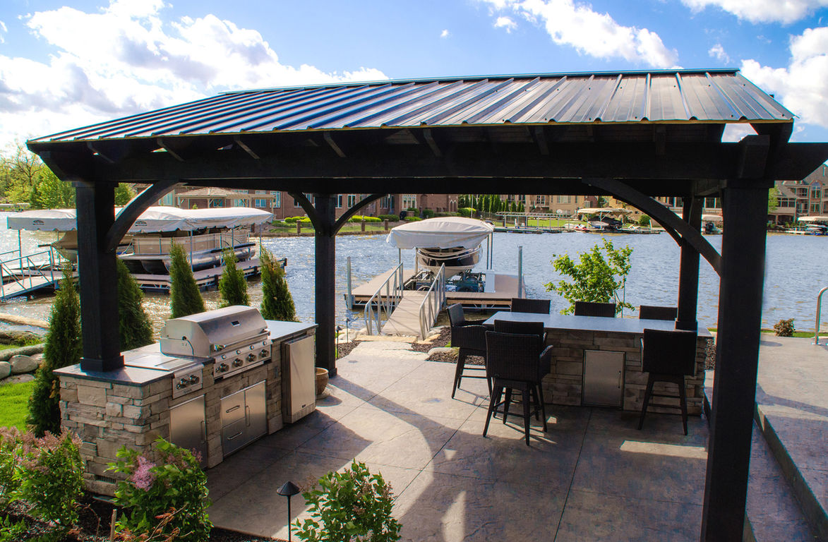 Lakeside pavilion with outdoor kitchen and grill beneath metal roof.