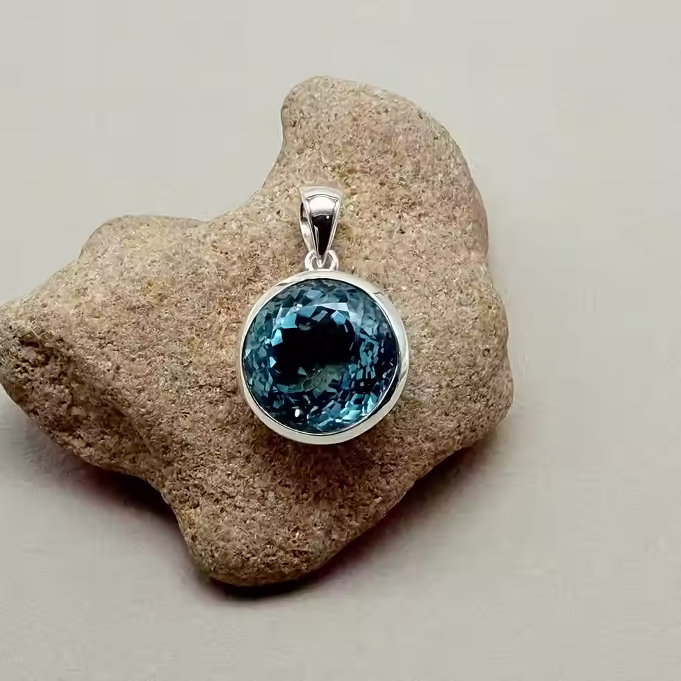 Sterling silver pendant featuring a round faceted blue topaz gemstone, displayed on a natural stone background.