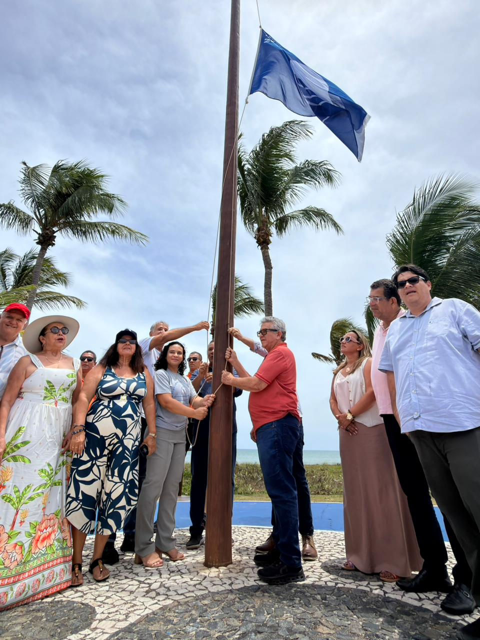 Hasteamento da Bandeira Azul 2025 na Praia do Paraíso, em Guarajuba, com autoridades, gestores e moradores presentes durante a cerimônia. A bandeira azul tremula ao lado do mar, simbolizando o selo internacional de qualidade ambiental conquistado pela praia.