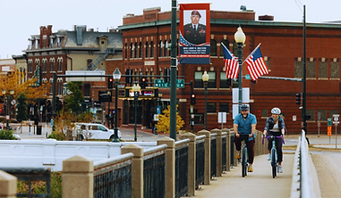 Cyclists on city bridge, American flags, military banner, historic brick buildings.
