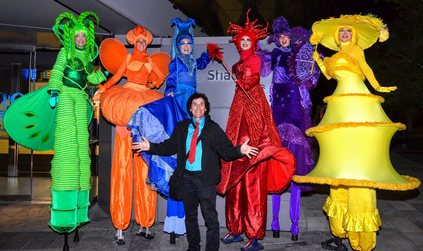 Un groupe d'artistes échassiers de Chromatika posant joyeusement avec un homme en costume, chacun portant des costumes brillants et colorés symbolisant les différentes couleurs de l'arc-en-ciel, prêts à animer un événement spectaculaire.