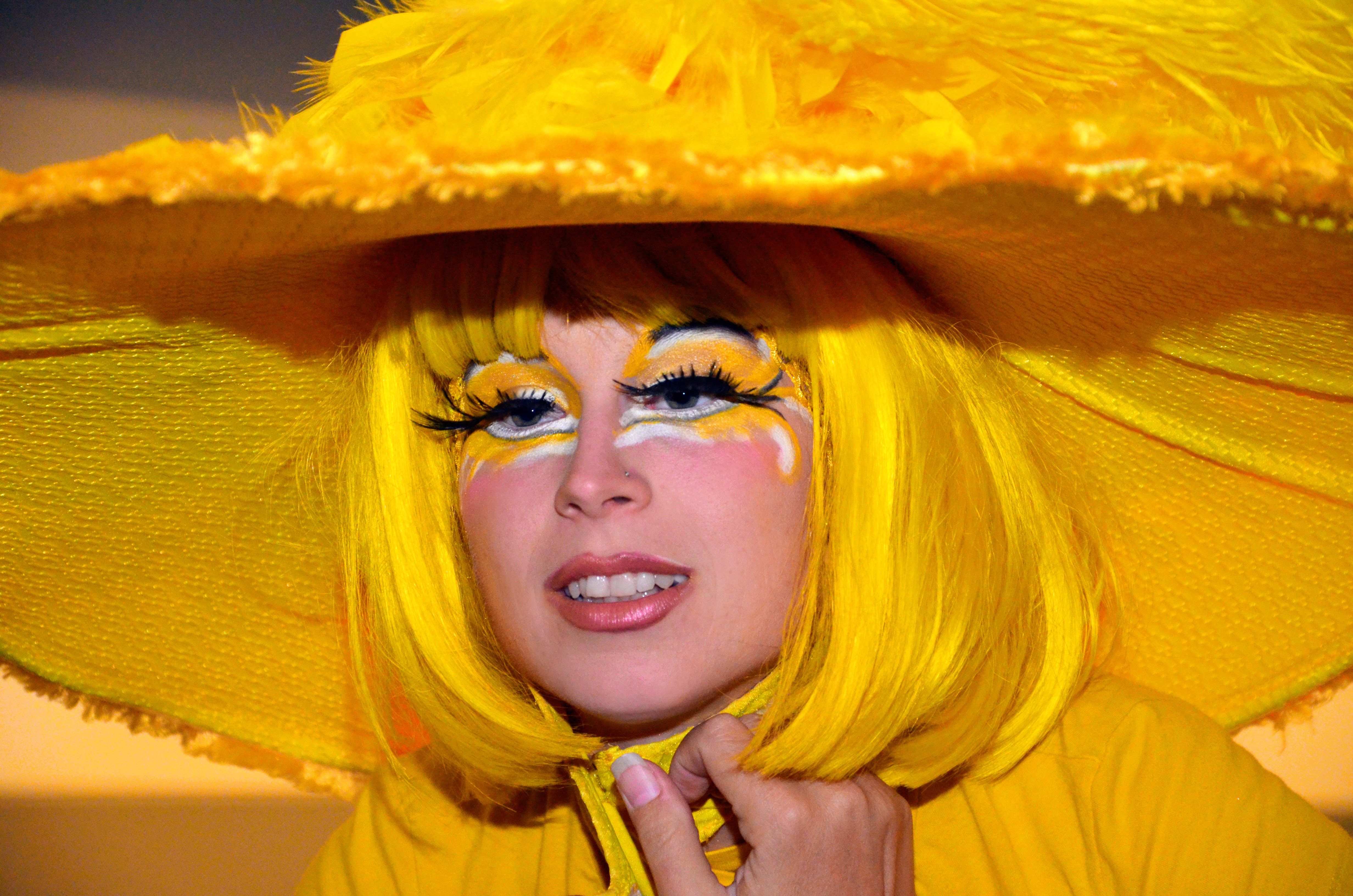 Artiste de cirque en costume jaune vibrant avec un grand chapeau assorti, maquillage théâtral mettant en valeur ses yeux expressifs, capturant l'émerveillement et le caractère spectaculaire d'une performance de rue.