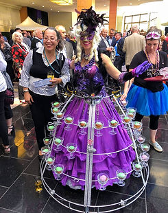 Artiste masquée en robe champagne violette et coiffe à plumes, entourée d’invités lors d’une réception.