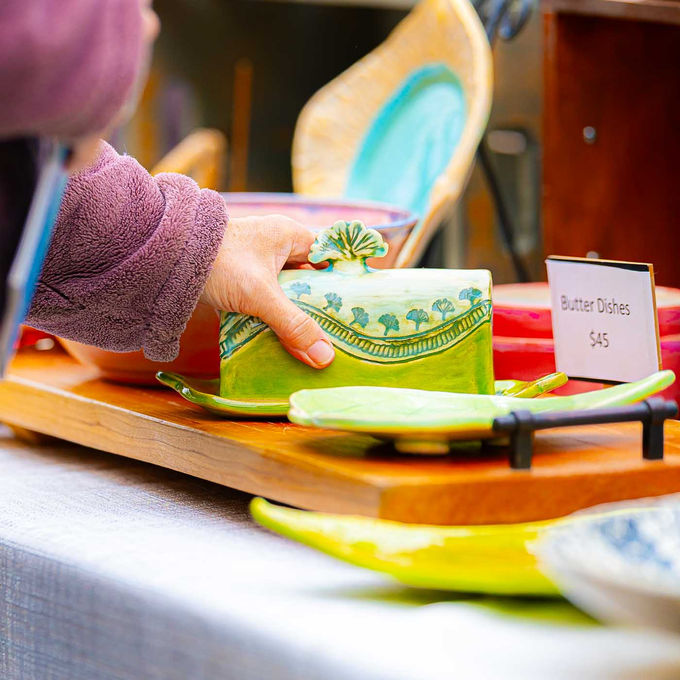 Hand lifting a green ceramic butter dish from a wooden display table at an art market.