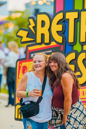 Two women posing and smiling at a colorful outdoor event sign, example of candid moments to look for when learning how to choose an event photographer