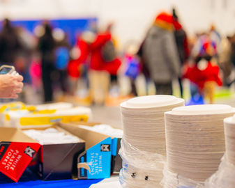Stacks of paper plates and serving supplies set up on a table inside a busy community event space, with people gathered in the background.