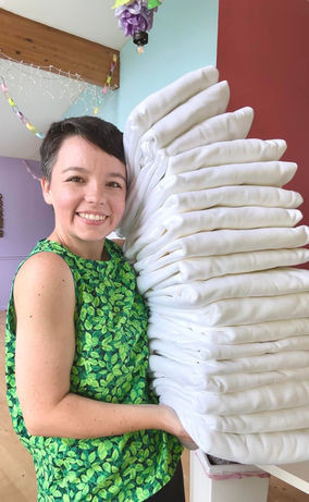Corky Lorenz is holding a tall stack of white sweatshirts in a colorful living room before they have been tie-dyed.