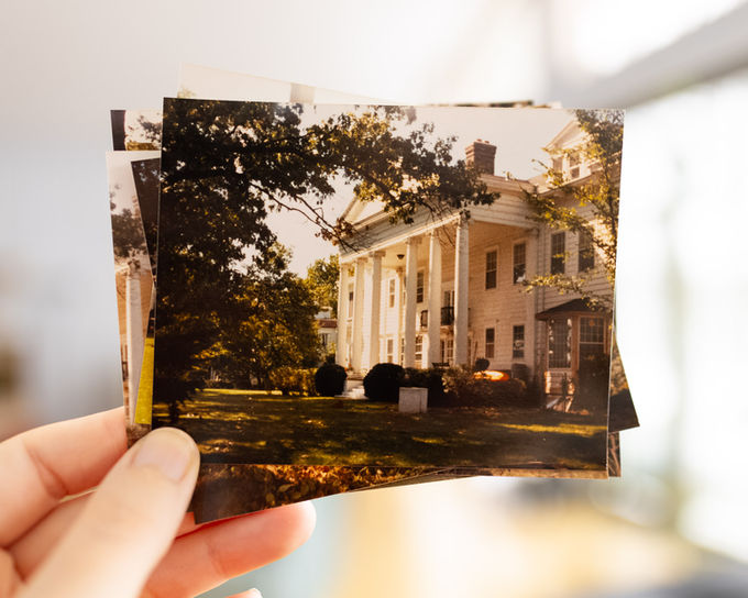 Hand is holding a stack of color, vintage photos of the Greenleaf mansion with windows in the background