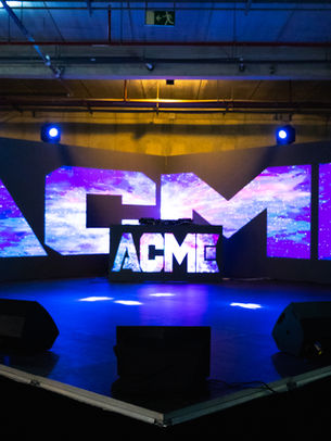 Escenario de evento con paredes de pantallas LED curvadas mostrando el logo de ACME en gráficos espaciales galácticos púrpuras y azules, cabina de DJ central, altavoces de suelo y focos de iluminación azules y amarillos en un almacén.
