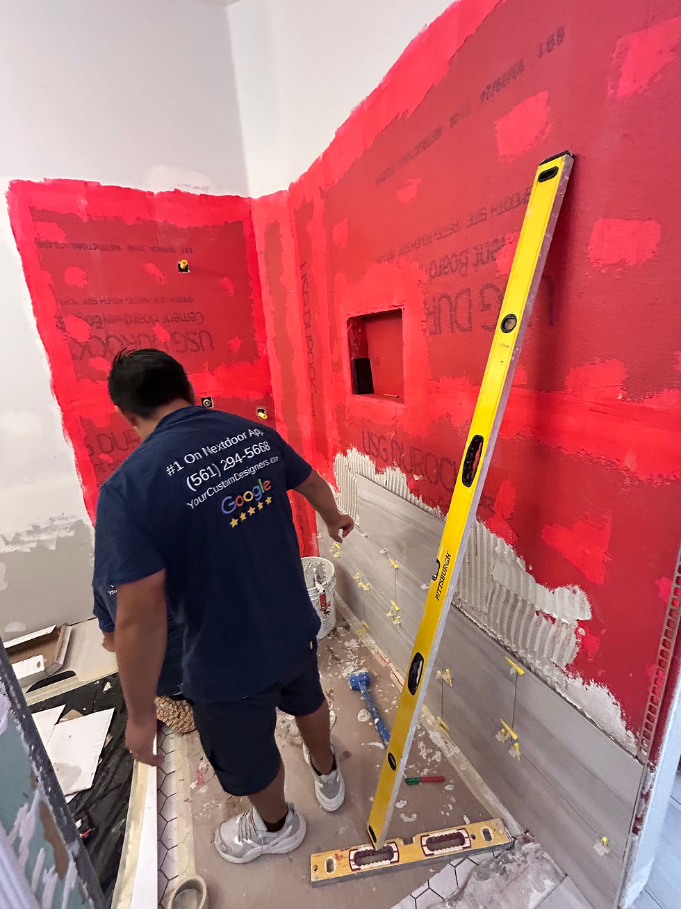 bathroom remodel with worker wearing blue Love By Design shirt and he is installing tile on a wall that is red from the waterproof coating.