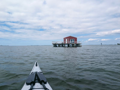 a kayak bow is in the foreground as it floats on a body of water with a red fish house on stilts is in the background