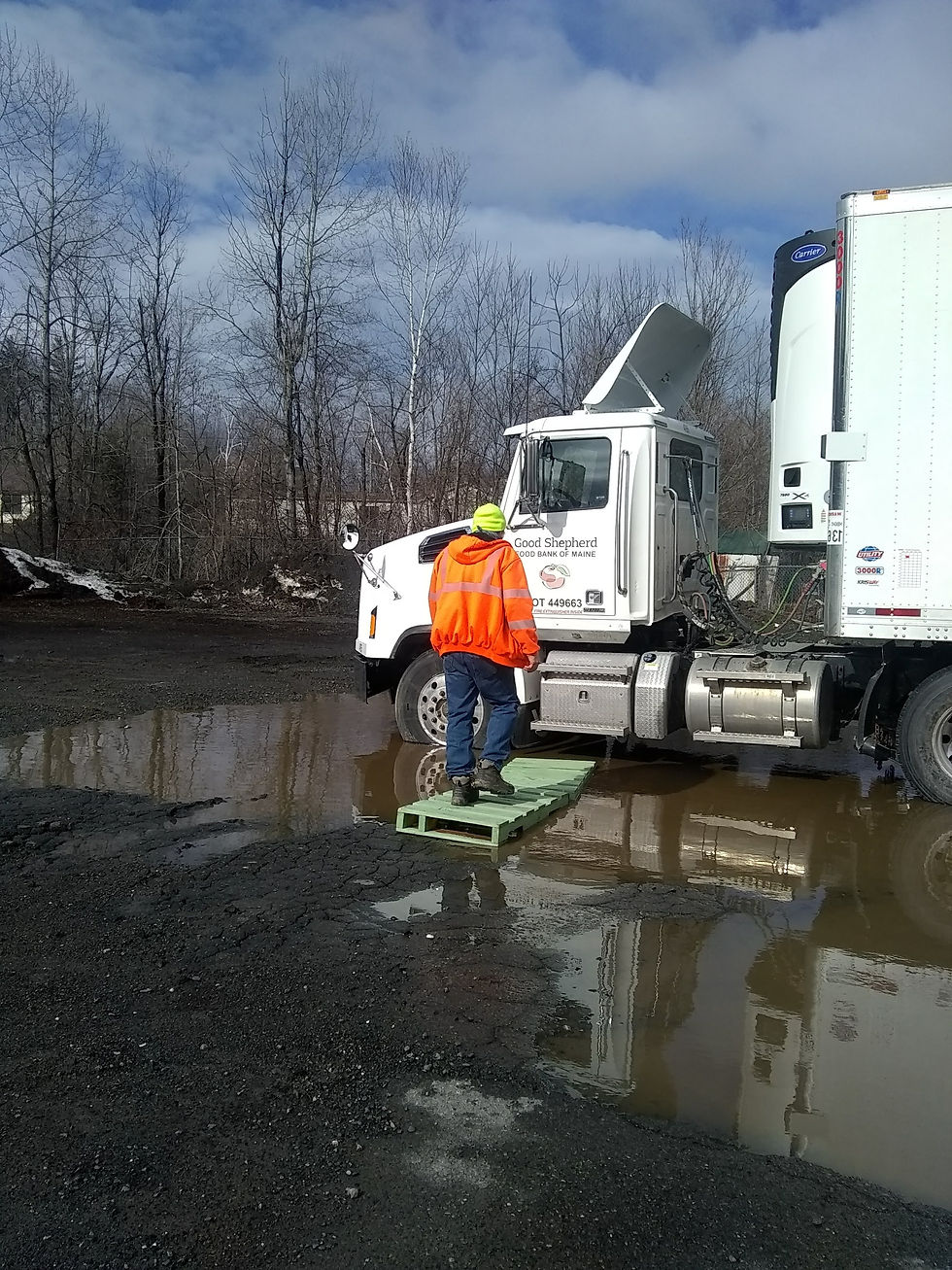A Good Shepherd delivery truck is parked in a deep puddle. The driver is walking across some wood planks to avoid getting soaked!