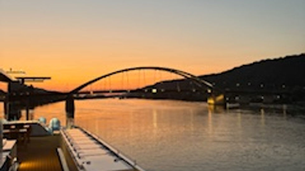 Sunset over a river with an arched bridge; view from a ship's deck. Sky transitions from orange to blue, creating a calm, serene mood.