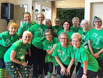 Thirteen women in green shirts huddle together with enthusiastic, joyful smiles on their faces.