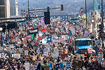 A crowd of protesters march holding signs and flags. Signs carry messages such as "Resist", "No Kings", and messages against military action in Iran.