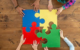 A square shaped puzzle consisting of four large pieces. Four pairs of hands collaborate to complete it. One pair is fitting in a large piece in the lower left hand corner.