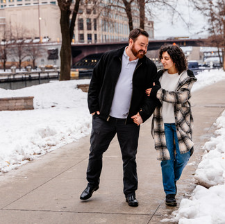A couple walks hand-in-hand on a snowy Chicago Riverwalk path, surrounded by leafless trees and buildings. They appear happy and relaxed. Chicagoland Wedding Photographer, Chicagoland Proposal Photographer, Chicagoland Engagement Photographer, Geneva Wedding Photographer, Chicago Riverwalk engagement session, snowy chicago engagement session, winter chicago engagement session