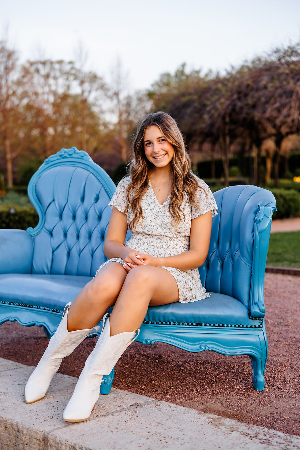 Smiling woman in white dress and boots sits on blue vintage sofa outdoors. best senior portrait photographers, family photographer, Geneva IL senior portraits, Wheaton senior portraits, west suburbs senior portrait photographers