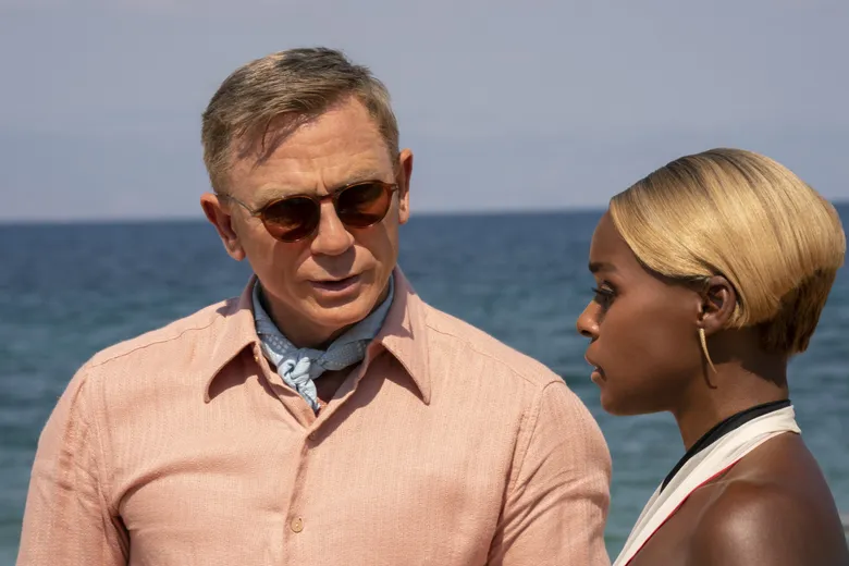 An still of Daniel Craig and Janelle Monae as Benoit Blanc and Helen Brand in the film Glass Onion. Craig stands on the left facing toward the camera while Monae stands to the right perpendicular to Craig, leaving only her side profile visible. Craig wears salmon pink button down and a light blue cravat. Monae's hair is blonde with brown roots and cut into a short bob. In the background behind them both rests only the sea and sky.