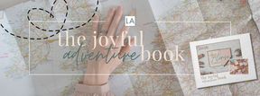 Hand on a world map with a marked route. Overlay text reads "the joyful adventure book." A photo of a family and the year 2026 are visible.