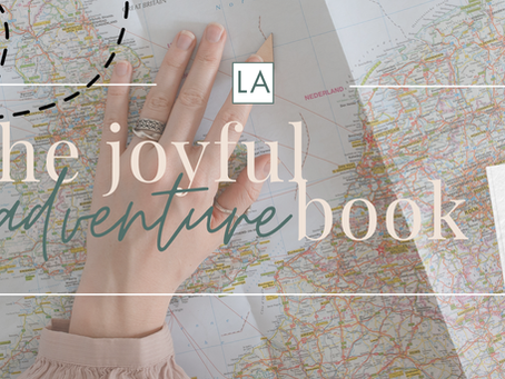 Hand on a world map with a marked route. Overlay text reads "the joyful adventure book." A photo of a family and the year 2026 are visible.