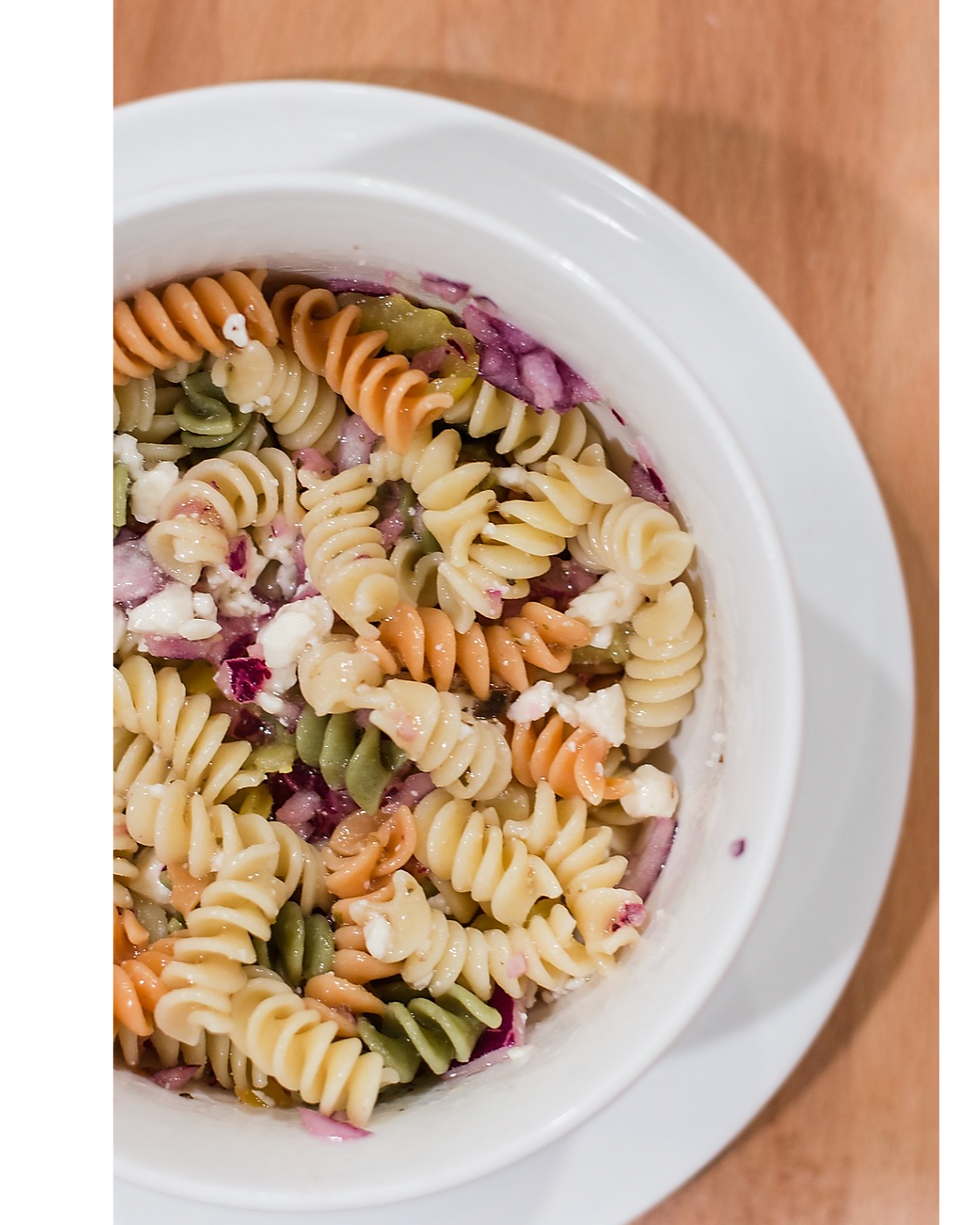 Tri-color rotini pasta salad with feta and onions in a white bowl on a wooden table, creating a vibrant and fresh look. greek pasta salad, holiday pasta salad, thanksgiving side dish, fresh holiday side, easy pasta salad, mediterranean pasta salad, christmas side dish