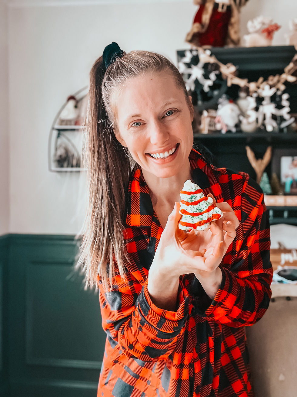 Smiling woman in a red plaid shirt holds a festive, decorated cupcake indoors. Background shows holiday decor and shelf with garland. homemade Christmas tree cakes, copycat Little Debbie Christmas tree cake, Christmas tree cake recipe from scratch, vanilla sheet cake Christmas dessert, marshmallow cream filling cake, white chocolate coated Christmas tree cakes, homemade holiday snack cake, nostalgic Christmas desserts, Christmas baking with kids, best Christmas tree cake recipe