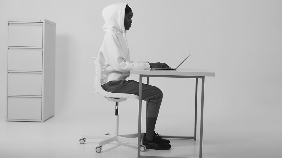 Person in a white hoodie sits at a desk using a laptop in a minimalistic, monochrome room. Chained to their desk with work.