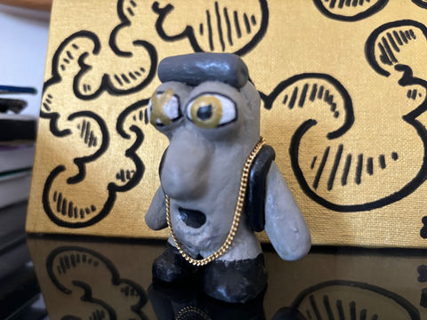 A small, hand-sculpted clay figure with a textured, grayish face, wide yellow eyes with black pupils, and white rings around them. It has a prominent nose and an open, circular mouth. The figure wears a dark gray flat cap and a thin gold chain around its neck. Its arms are short and stubby, and it wears black shoes. The sculpture stands on a reflective black surface in front of a gold canvas with a repeating black cloud pattern. The artist's signature 'E.P.o 2024' is visible on the canvas.