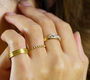 The model places their hand near their ear, wearing gold rings on each finger.