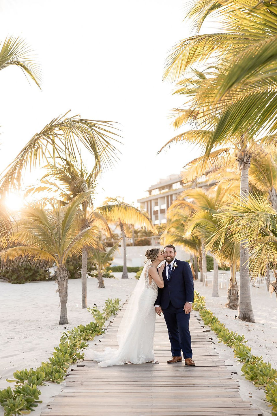 Bride kissing groom's cheek on beach boardwalk at Majestic Elegance Costa Mujeres.