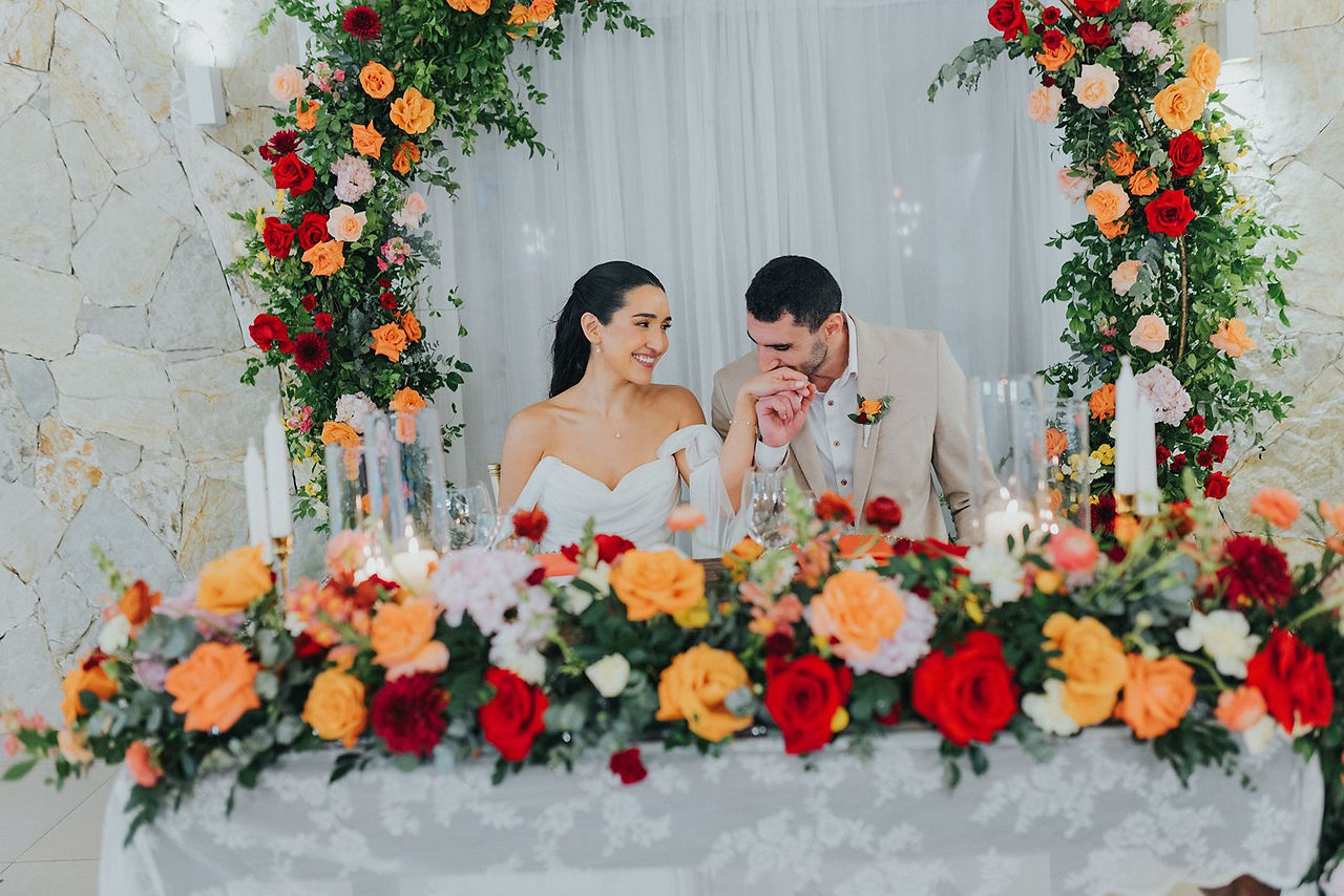 Groom kissing bride's hand at sweetheart table surrounded by coral and red flowers.