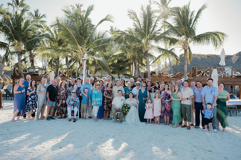 A large destination wedding group poses on a sunny beach with palm trees at Dreams Royal Beach resort in the Dominican Republic.
