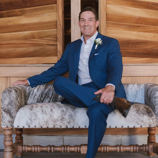 Groom sits confidently on a cowhide-upholstered bench wearing a navy blue suit with a white boutonniere, framed by a warm wood-paneled wall with a relaxed, polished look.