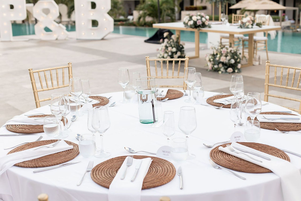 Simple and modern wedding tablescape at Sky Lounge venue at Majestic Elegance Costa Mujeres, featuring woven charger plates, white linens, gold chiavari chairs, and poolside floral decor.