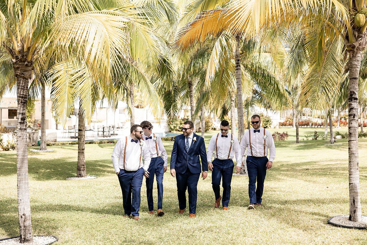 Groomsmen in white shirts, navy pants and brown suspenders walking with tropical background in Mexico.