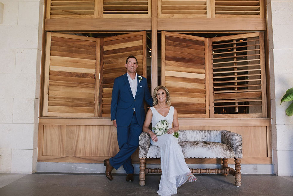 Bride sitting couch holding white bouquet and groom in navy blue suite leaning against a wooden wall.
